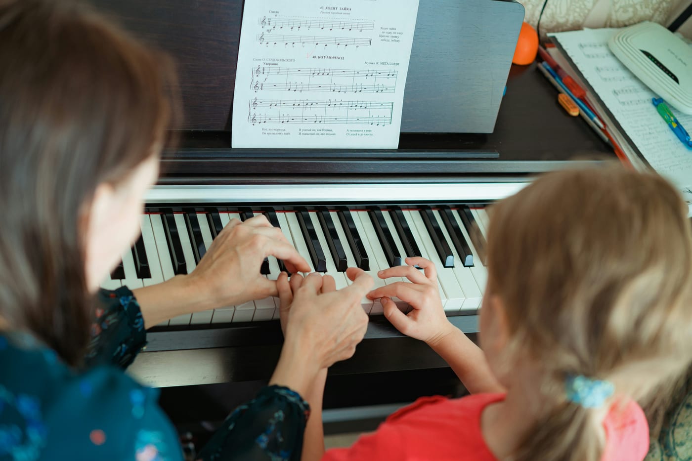 Teacher guiding a young student's hands on the piano during a lesson, sheet music open above the keys.